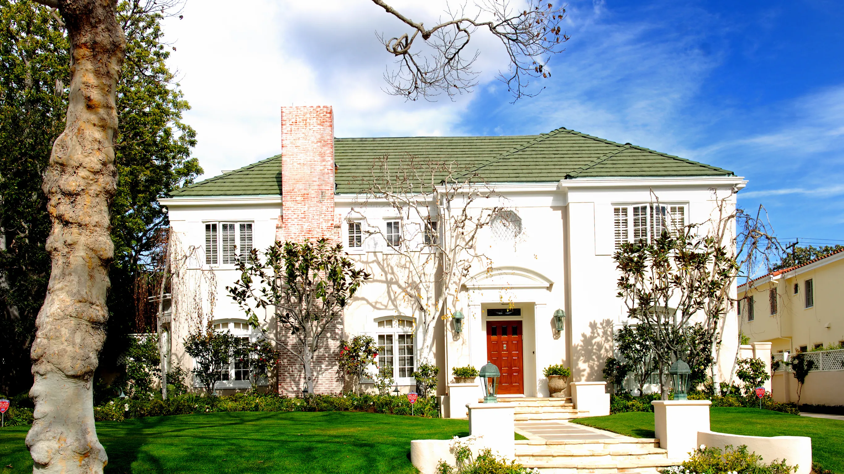 After home restoration project in Los Angeles showing a restored classic home exterior with white stucco walls, green roof, red front door, brick chimney, landscaped lawn, and finished curb appeal