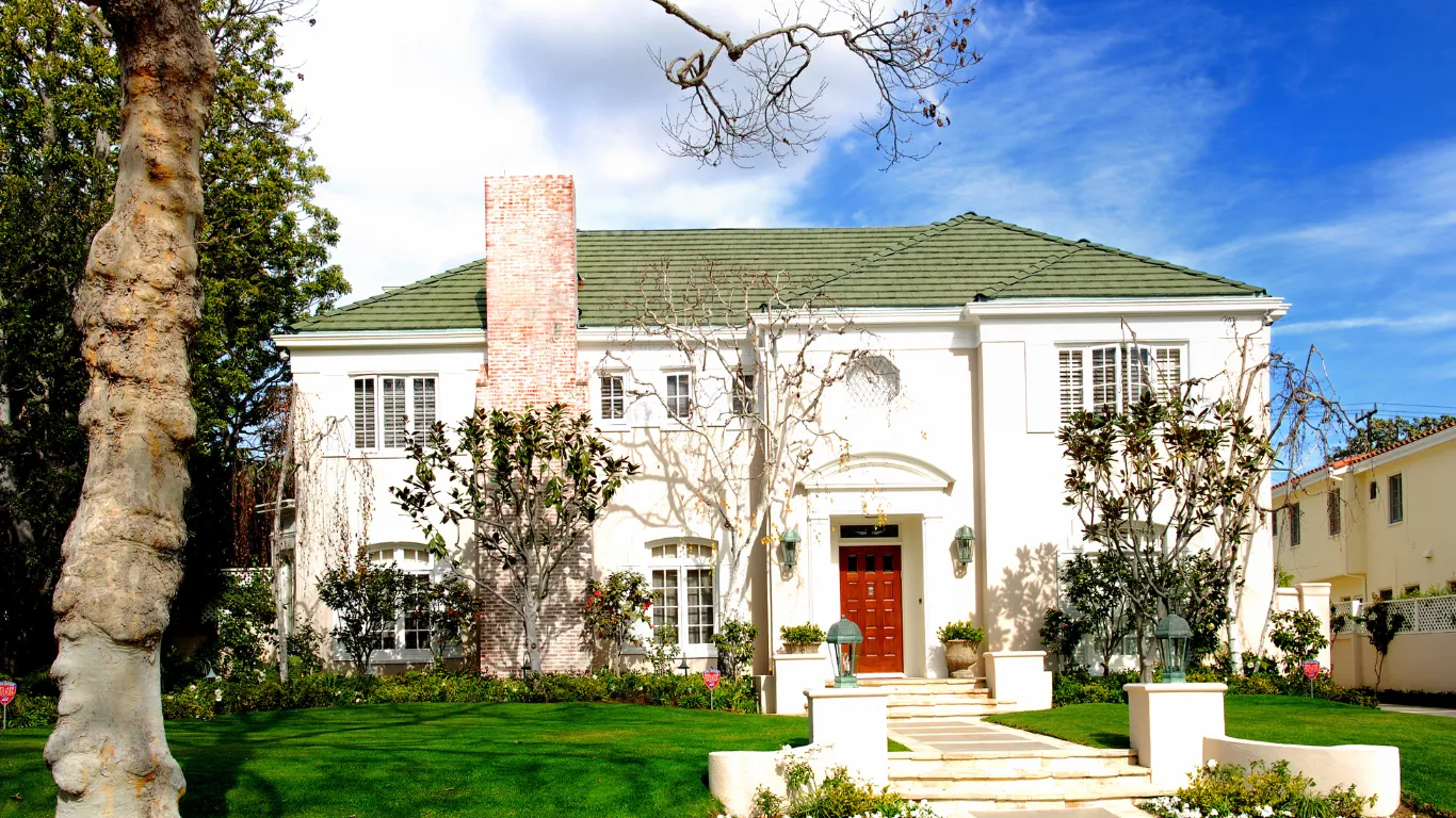 Home restoration project in Los Angeles showing a restored classic home exterior with white stucco walls, green roof, red front door, brick chimney, landscaped lawn, and finished curb appeal