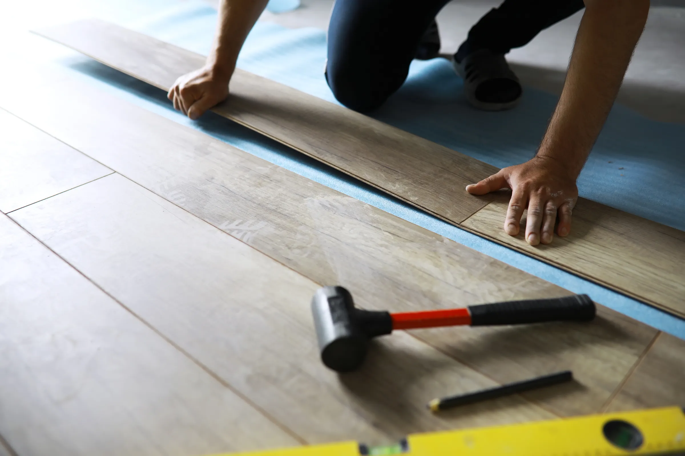 Flooring installation in Los Angeles showing laminate wood plank flooring being installed with underlayment, rubber mallet, pencil, and level tool